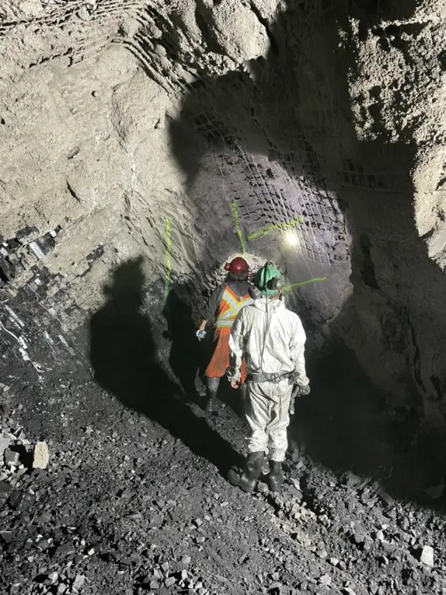 Two miners walking an underground drift, mesh and bolts on the walls and back